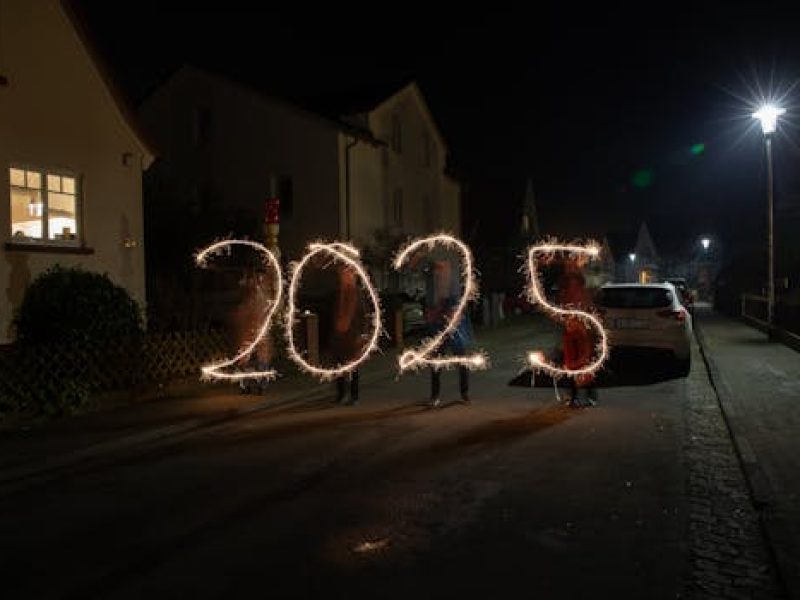 Sparkler light painting forming '2025' in a festive night street scene.
