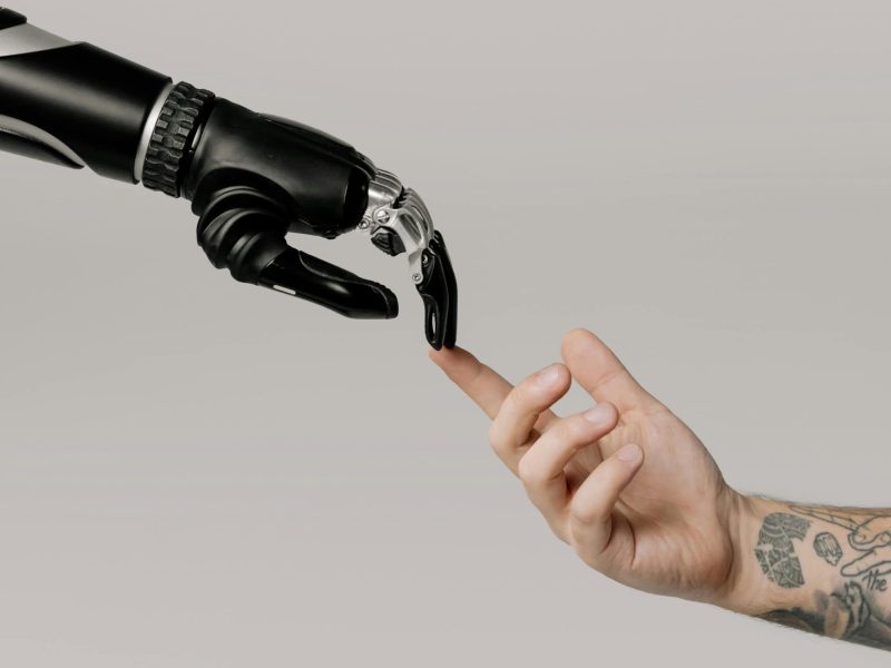 pexels-photo-6153354-6153354 A human hand with tattoos reaching out to a robotic hand on a white background.